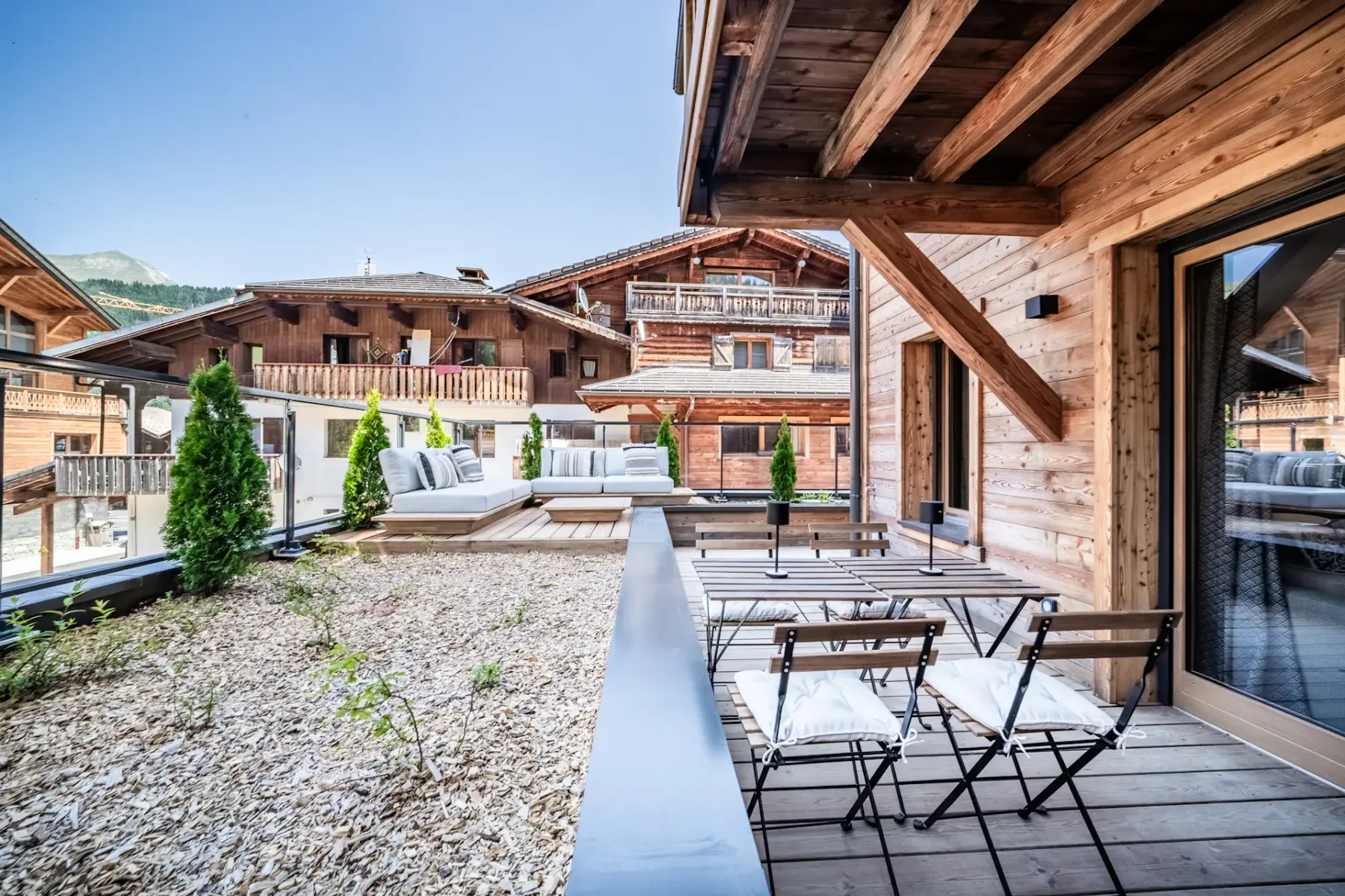 Una terraza de madera con asientos al aire libre da a un patio de grava y edificios cercanos.