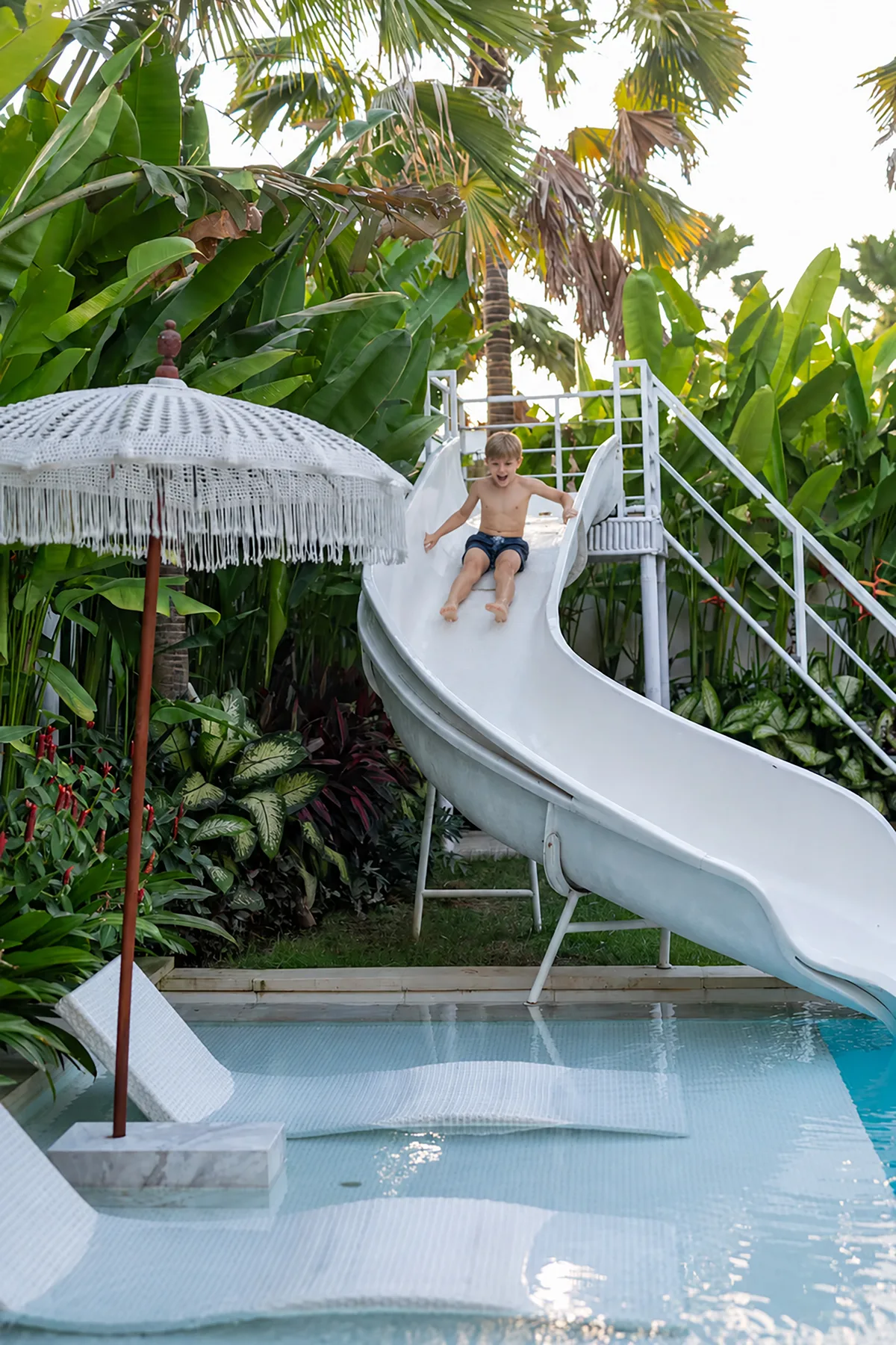 Un niño se desliza hacia una piscina rodeada de exuberantes plantas tropicales.