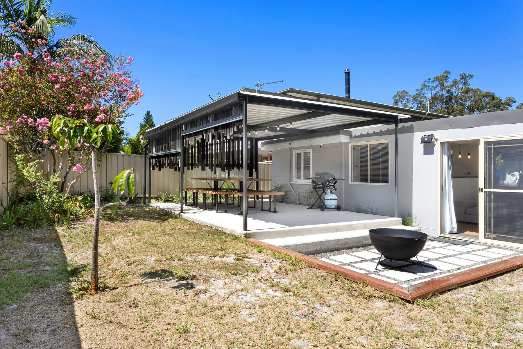 Un patio exterior cubierto junto a una casa, con una parrilla y jardín.