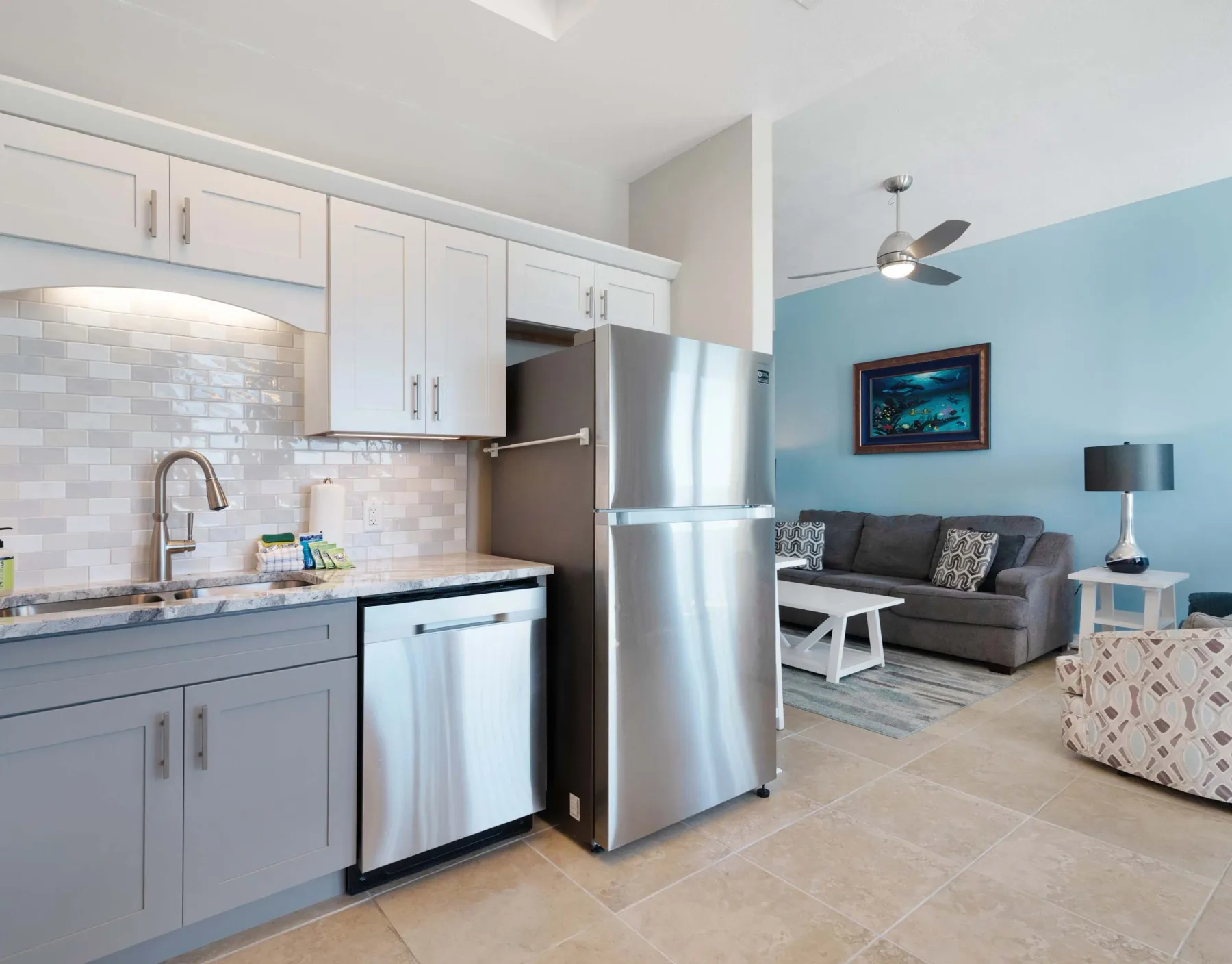 Cocina y sala de estar de concepto abierto con un sofá gris y un refrigerador de acero inoxidable.