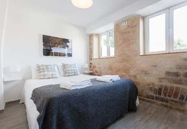 Un dormitorio luminoso con una cama, toallas dobladas y grandes ventanas.