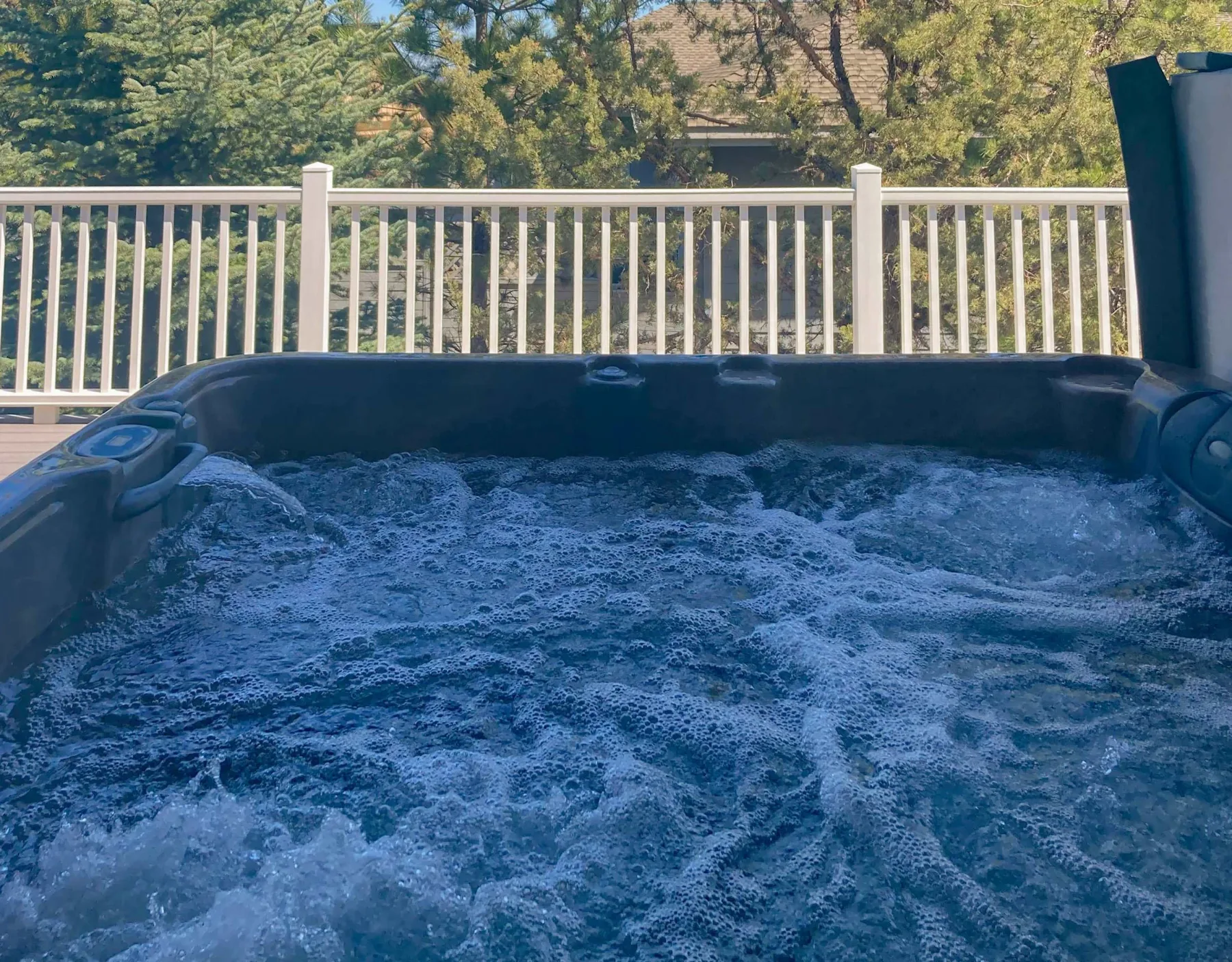 Una bañera de hidromasaje burbujeante está junto a una barandilla blanca, con árboles al fondo.