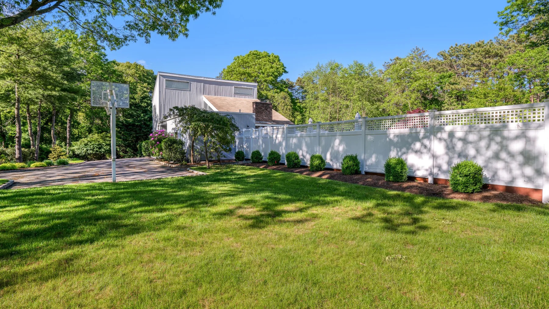 A grassy yard with a basketball hoop beside a house and white fence.