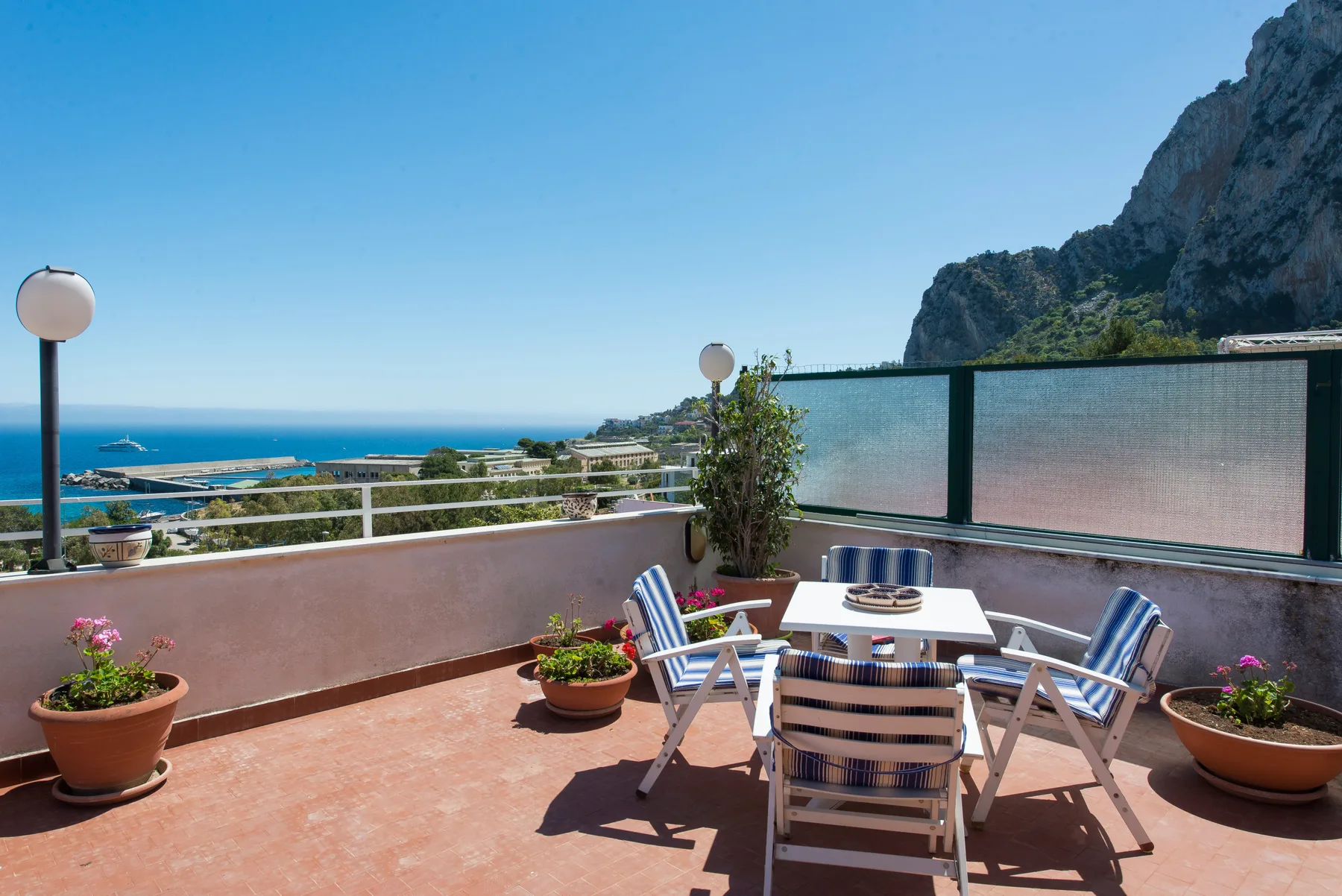 A rooftop terrace with outdoor seating overlooks the sea and a rocky hillside.