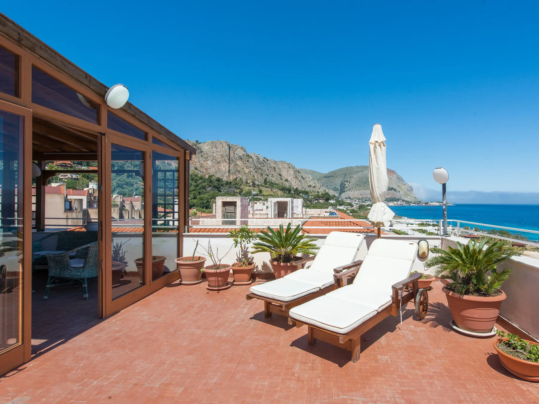 A sunny terrace with lounge seating, potted plants, and a view of the sea.