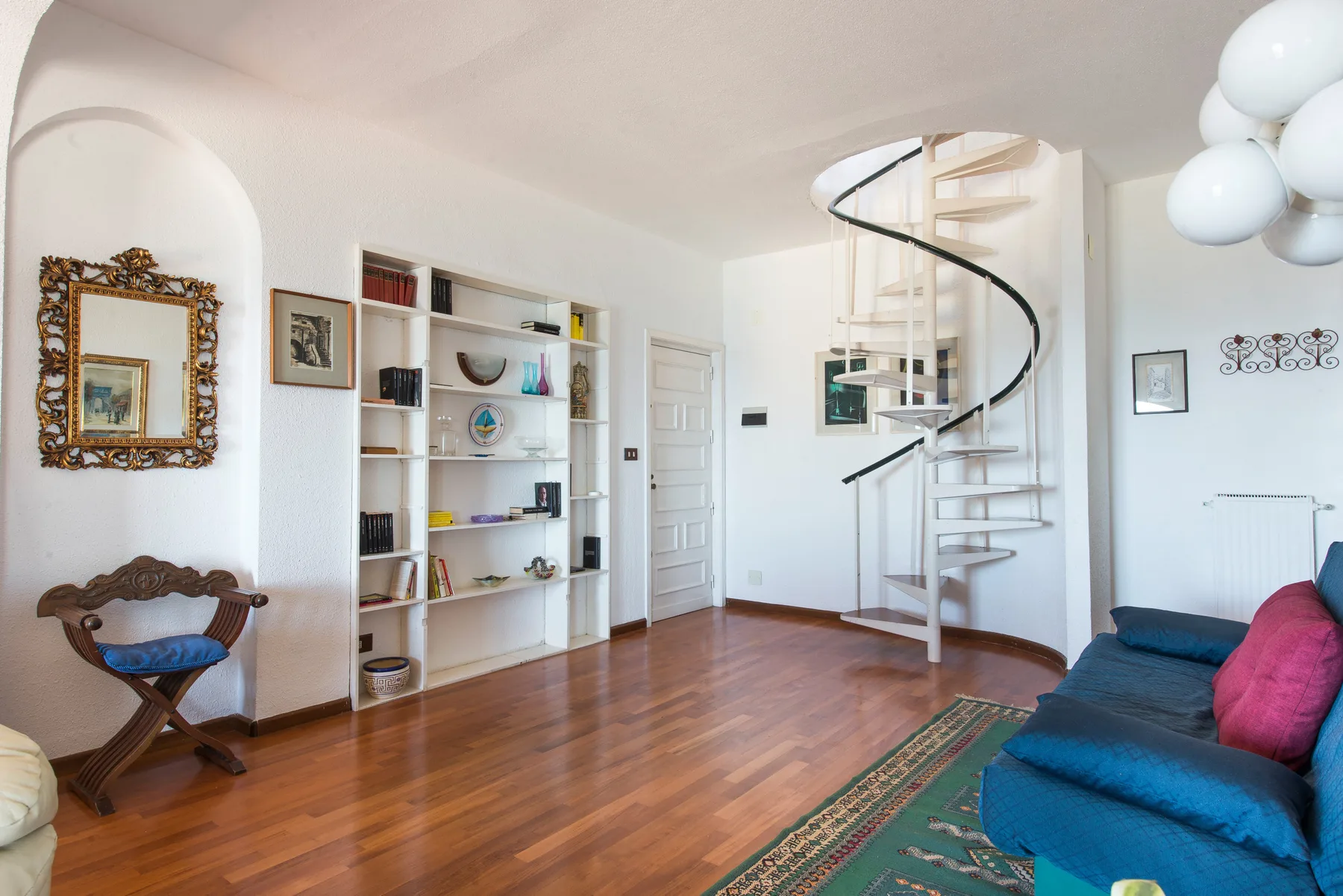 A bright living room with a spiral staircase, shelving, and blue seating.