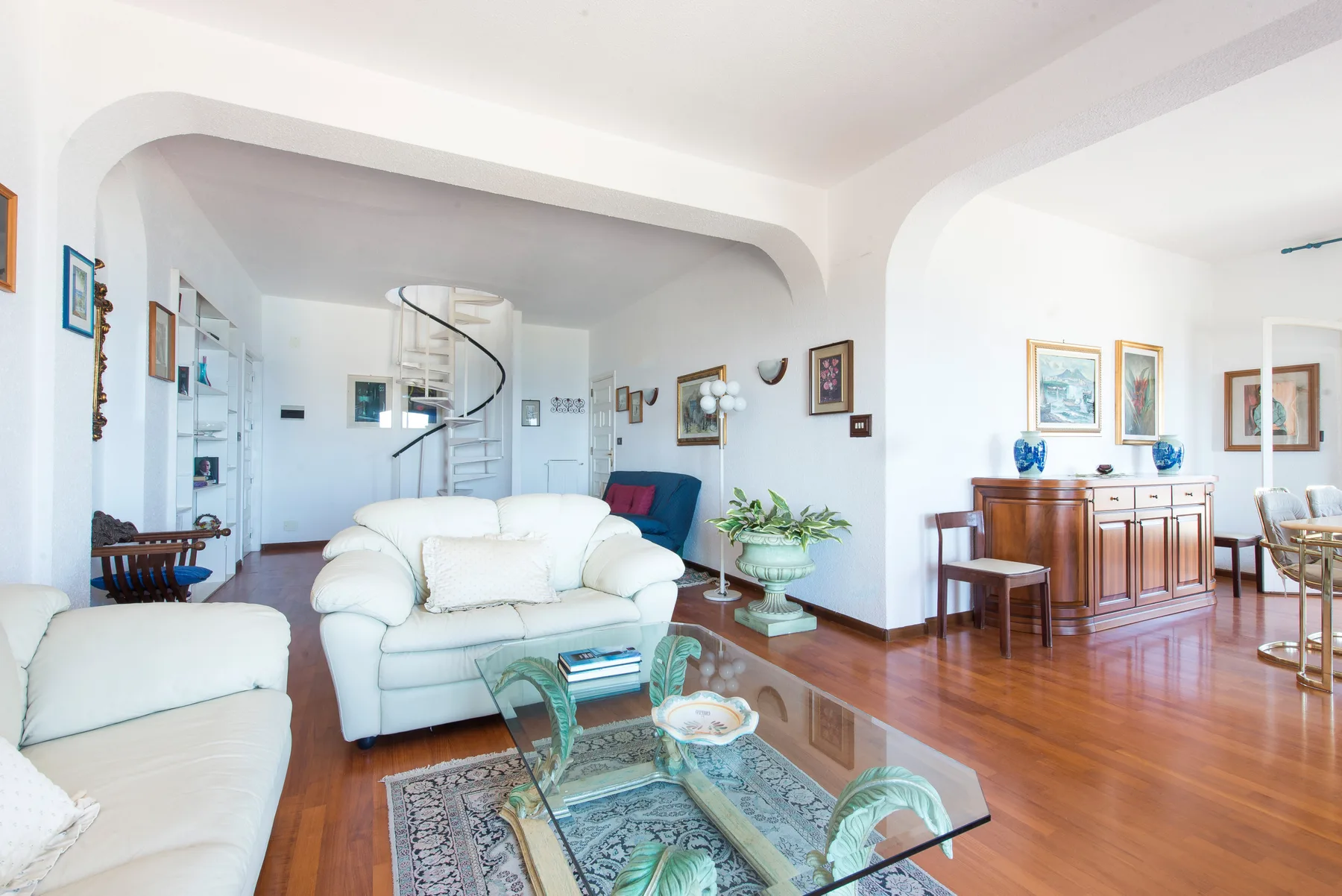 A bright living room with white seating, a glass coffee table, and a spiral staircase.