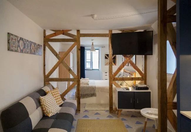 Una sala de estar con un sofá estampado y vigas de madera, abierta hacia un dormitorio.