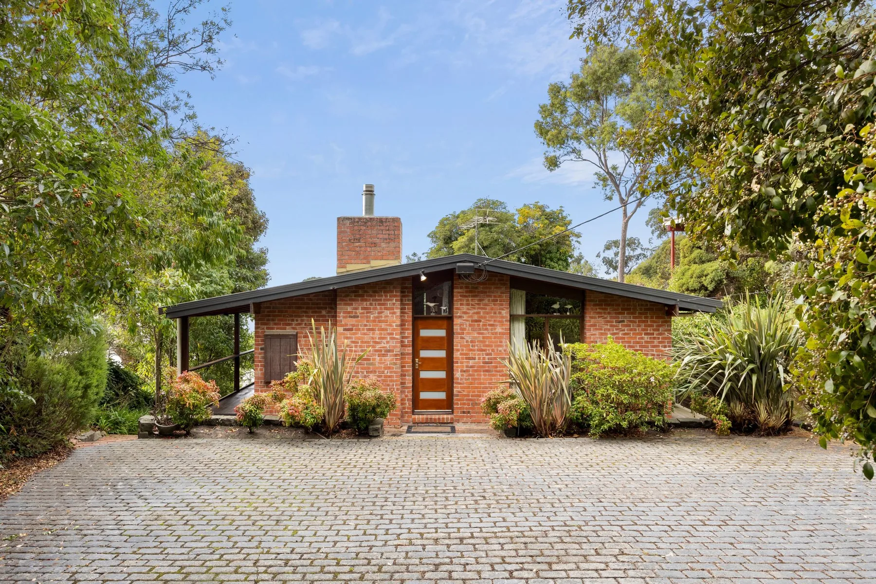 A single-story brick house with a wooden front door sits among leafy trees.
