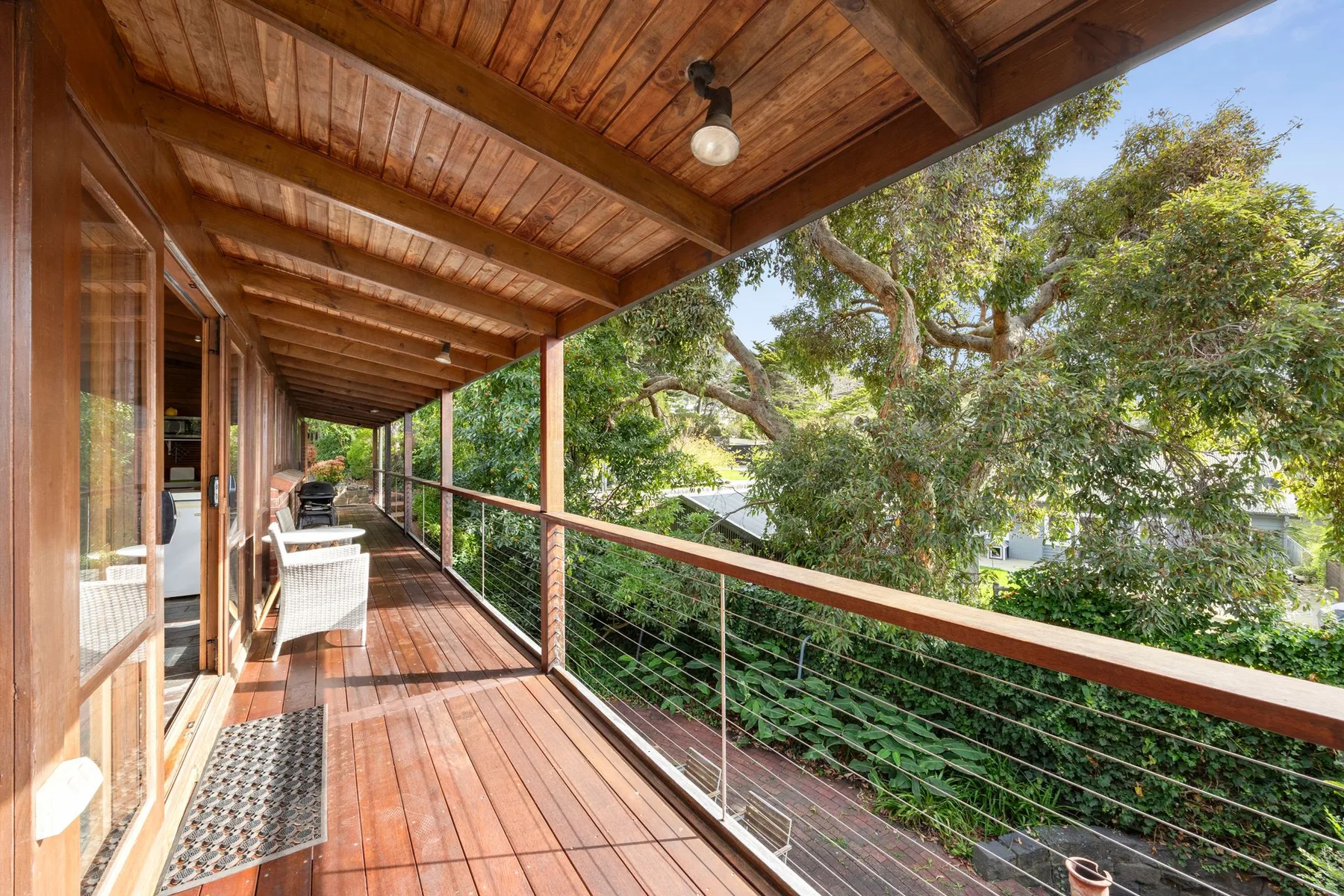 A covered wooden balcony runs alongside the house, overlooking leafy trees.