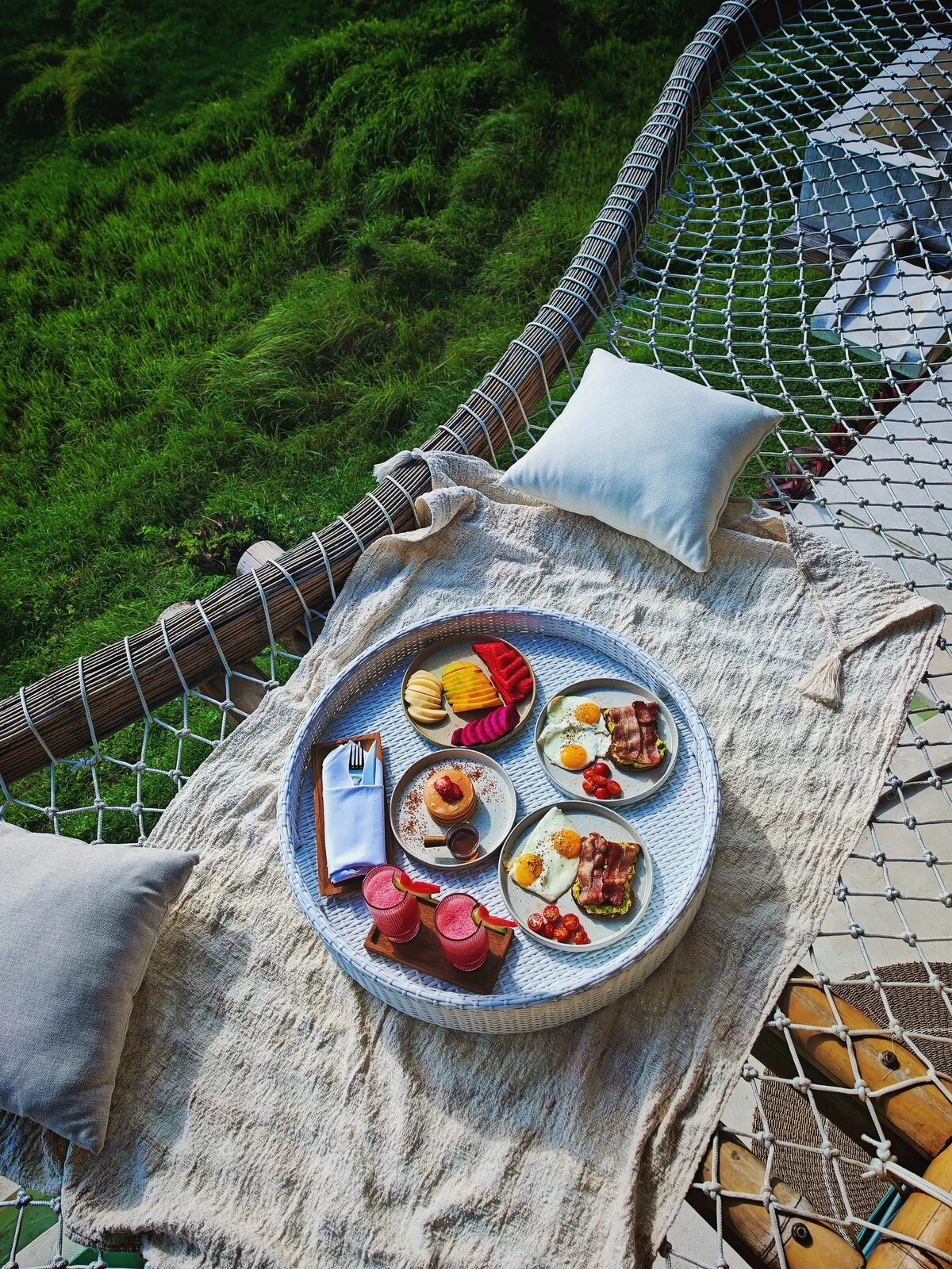 A breakfast tray with pastries and fruit rests on a blanket in an outdoor hammock.
