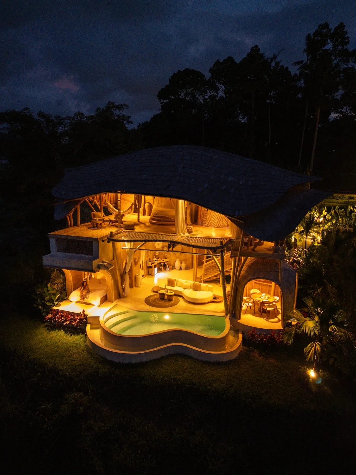 A warmly lit open-sided lodge sits beneath a broad roof amid dark trees.
