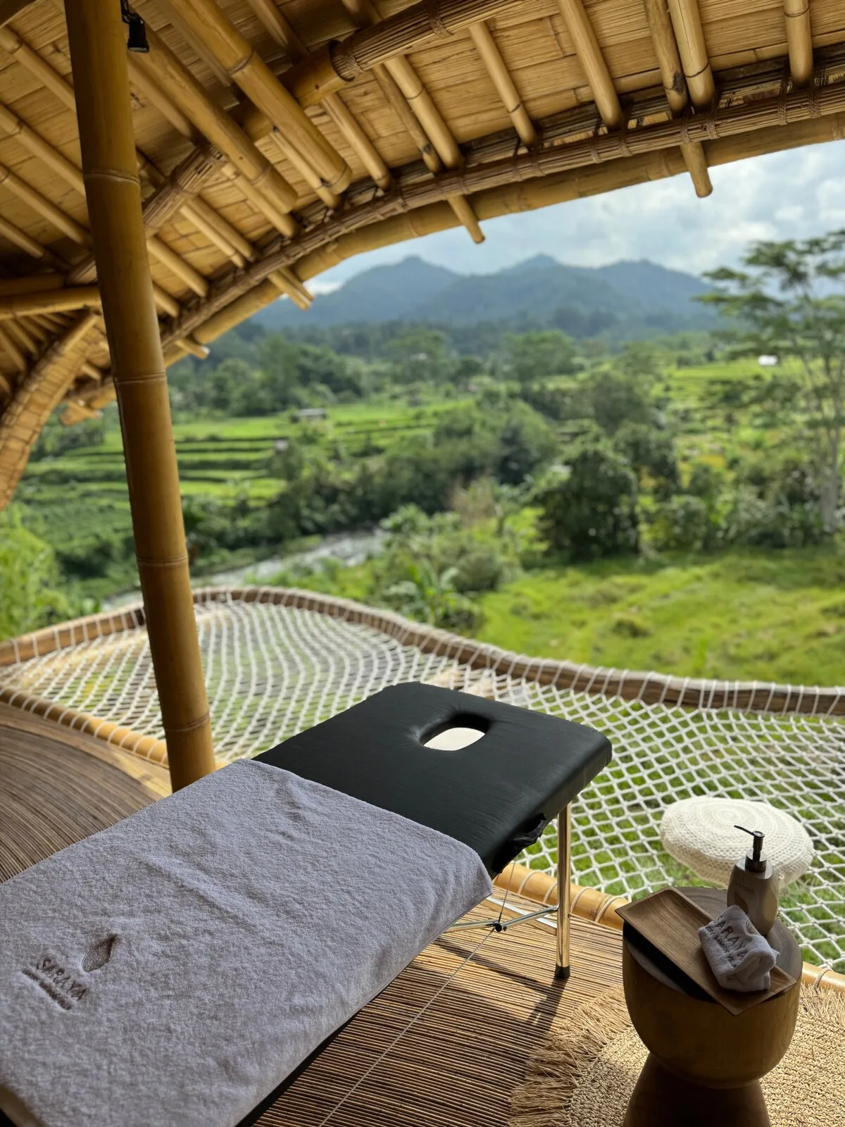 A massage table sits on a bamboo-covered outdoor terrace overlooking green hills.