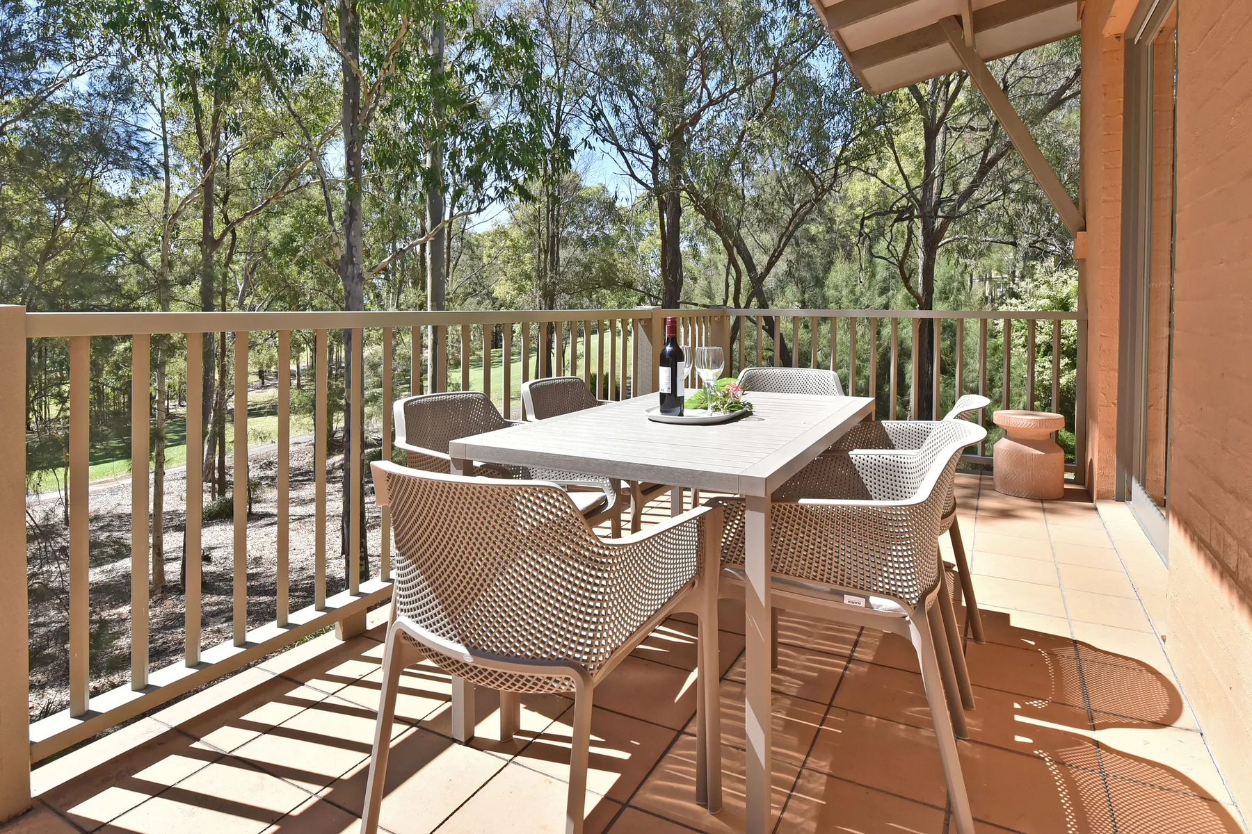A furnished outdoor dining area sits on a sunlit deck overlooking trees.