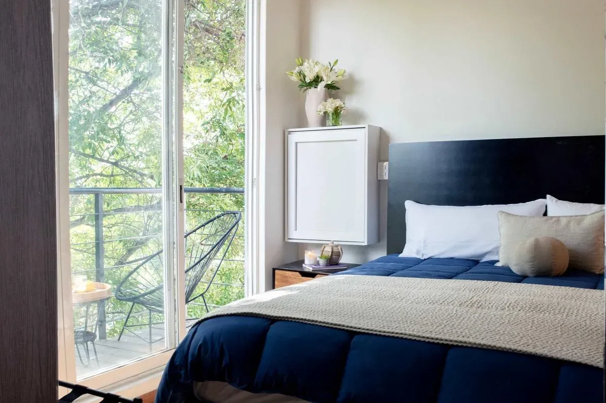A bedroom with a bed beside tall windows overlooking leafy trees.