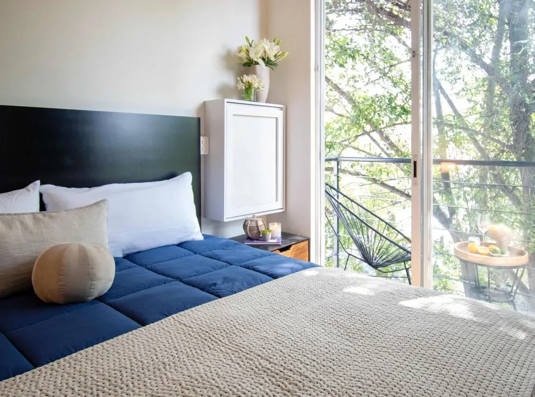 A bedroom with a bed beside large glass doors opening onto a leafy balcony.
