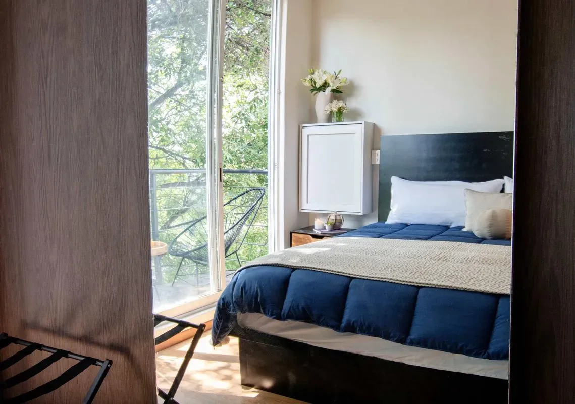 A bedroom with a bed beside tall windows overlooking leafy trees.