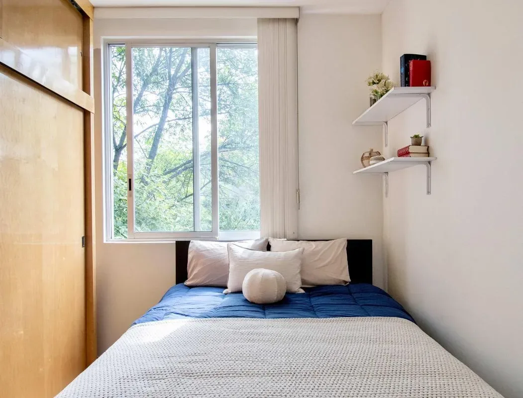 A bedroom with a bed beneath a window overlooking leafy trees.