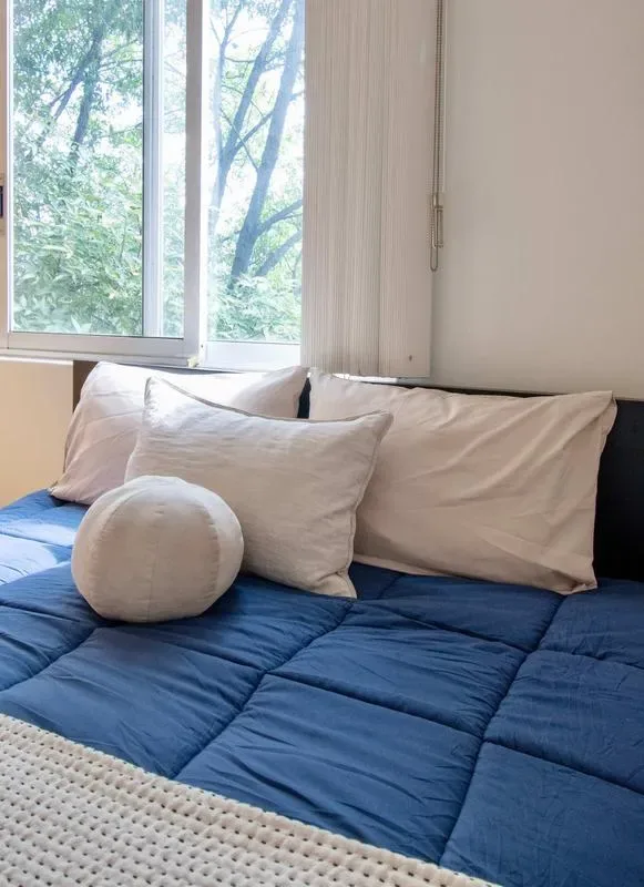 A bed with blue bedding and pillows sits beside a window overlooking trees.