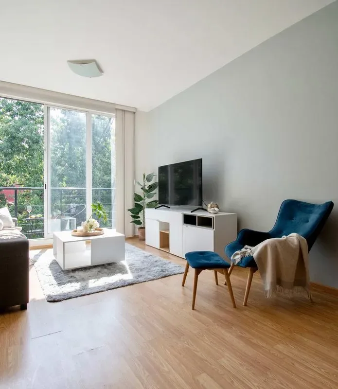 A bright living room has a television, seating, and large windows overlooking greenery.