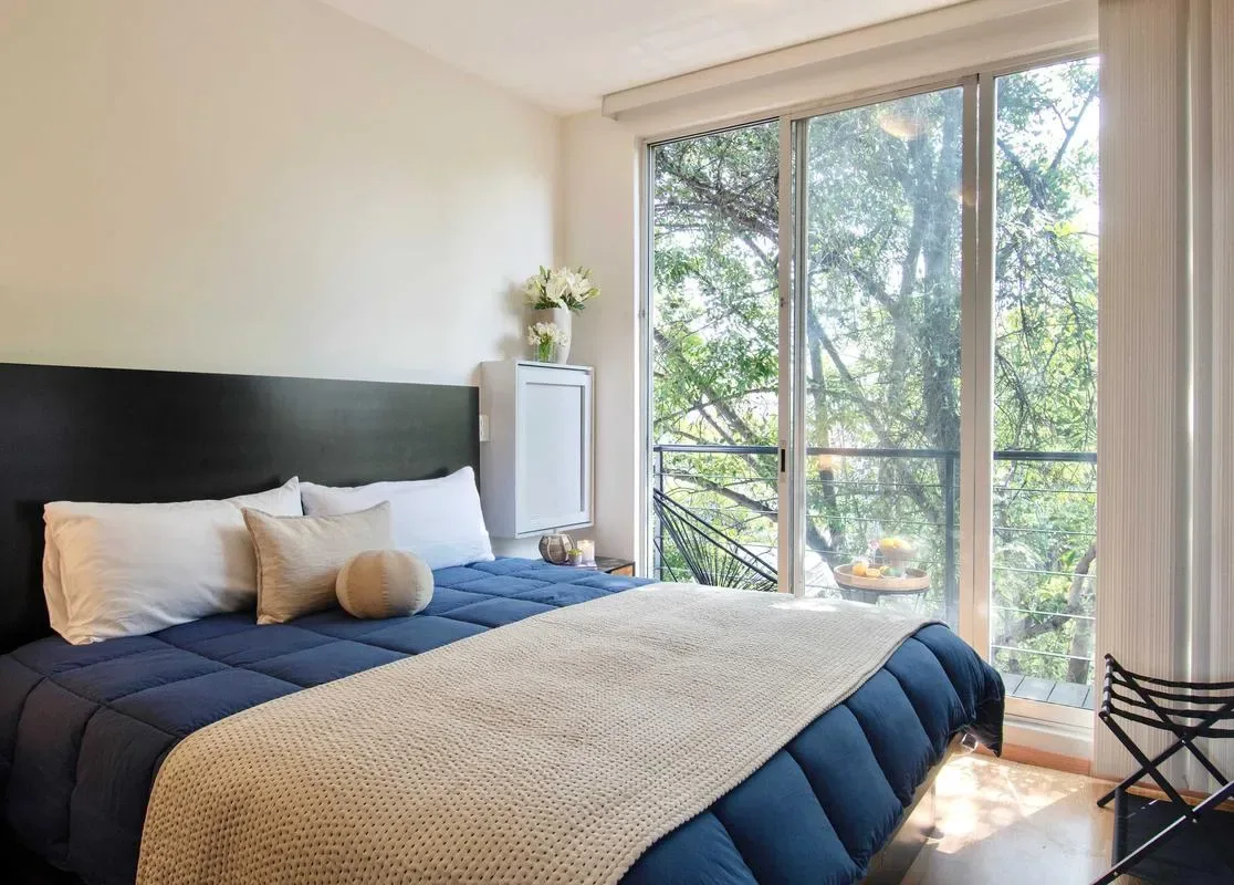 A bedroom with a blue bed beside large windows overlooking leafy trees.