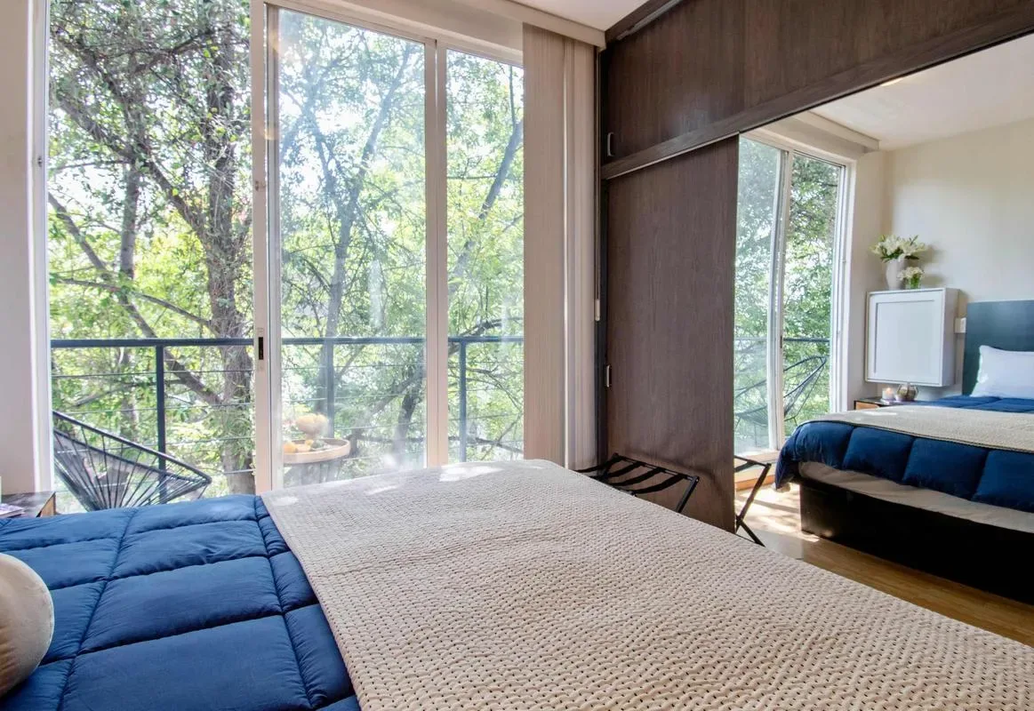 A bedroom with a bed beside large windows overlooking leafy trees.
