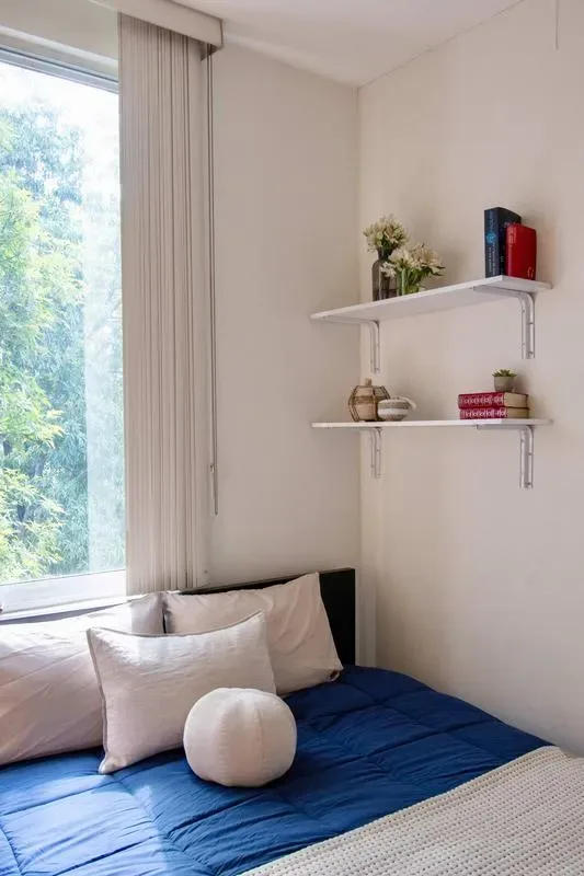 A bedroom with a blue bed beside a large window and wall shelves.
