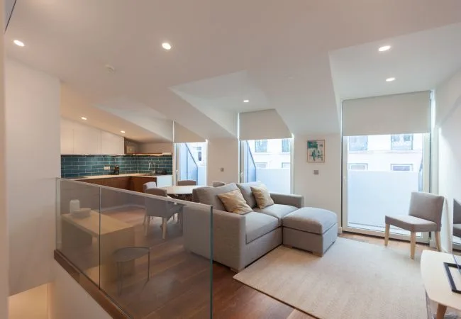 Una sala luminosa con sofá seccional, comedor y ventanas amplias.