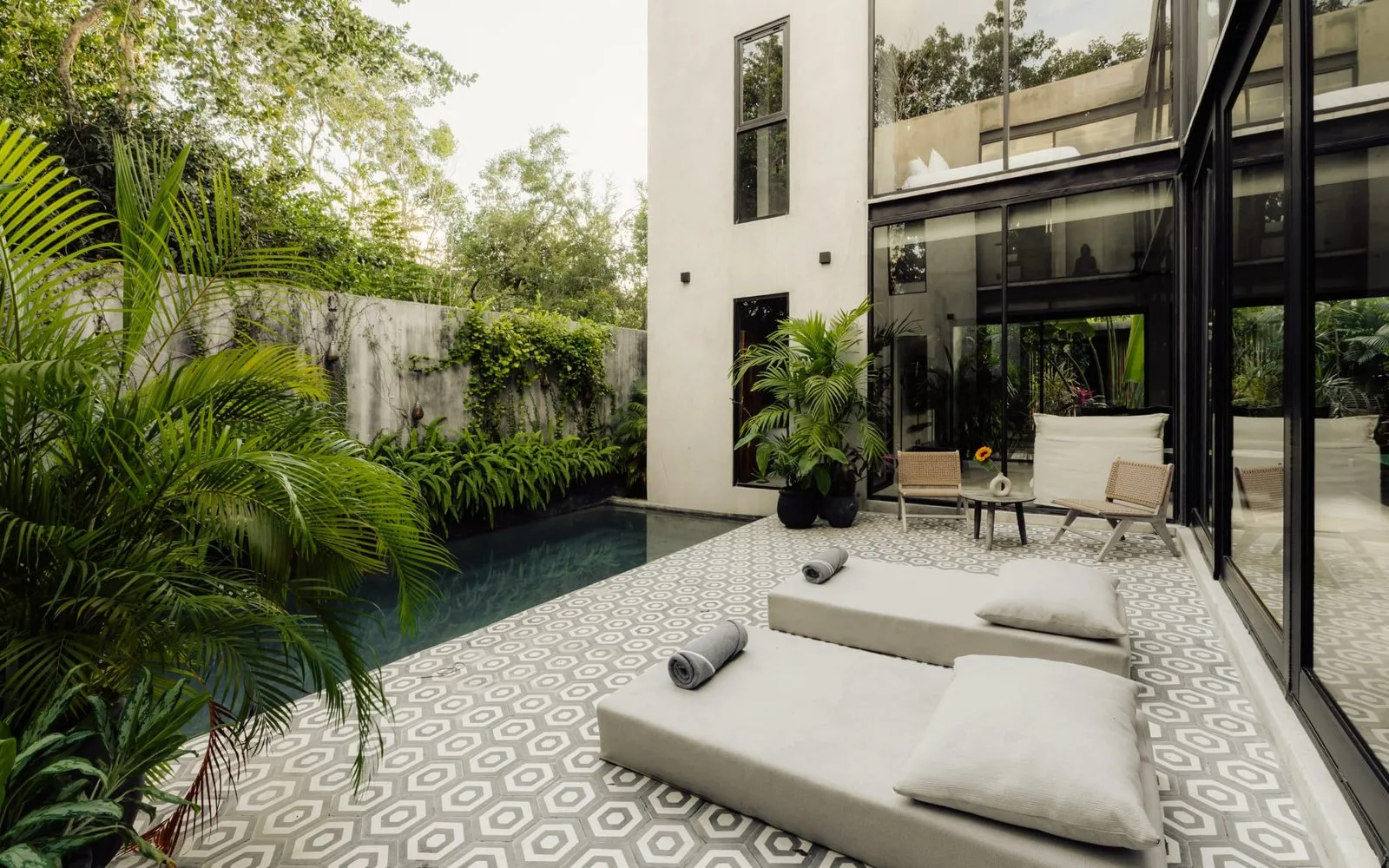 A tiled patio beside a narrow pool, with cushioned loungers and leafy plants.