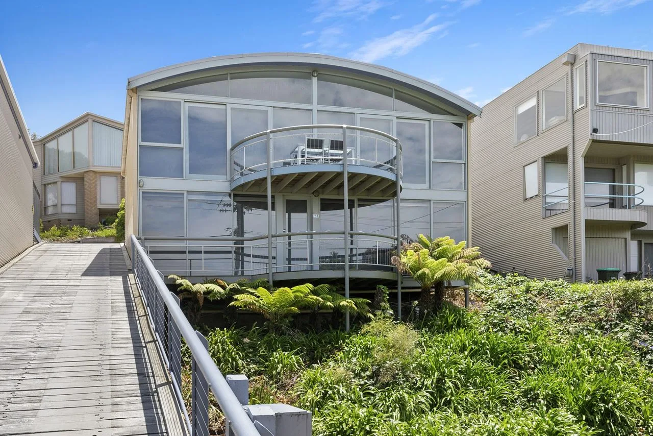 Un edificio moderno con fachada de vidrio se alza sobre un jardín junto a una pasarela elevada.
