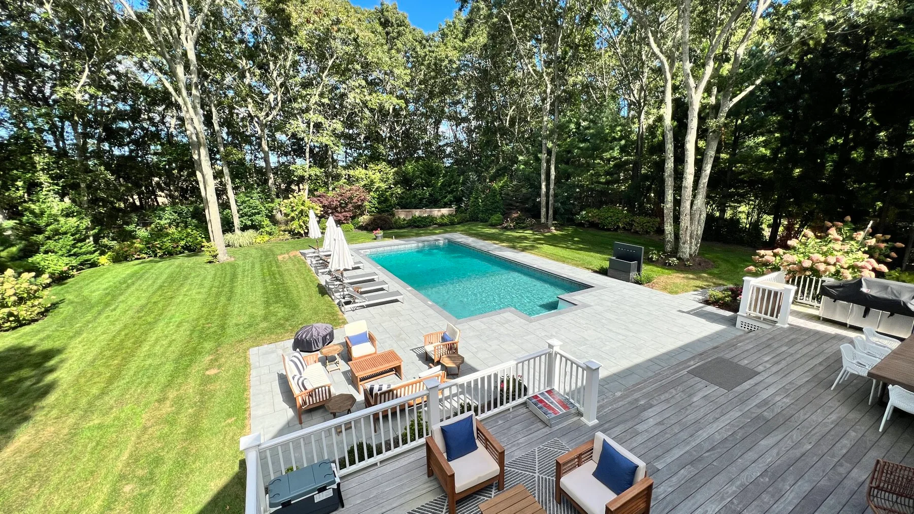 An outdoor swimming pool beside a deck, patio seating, and a green lawn.