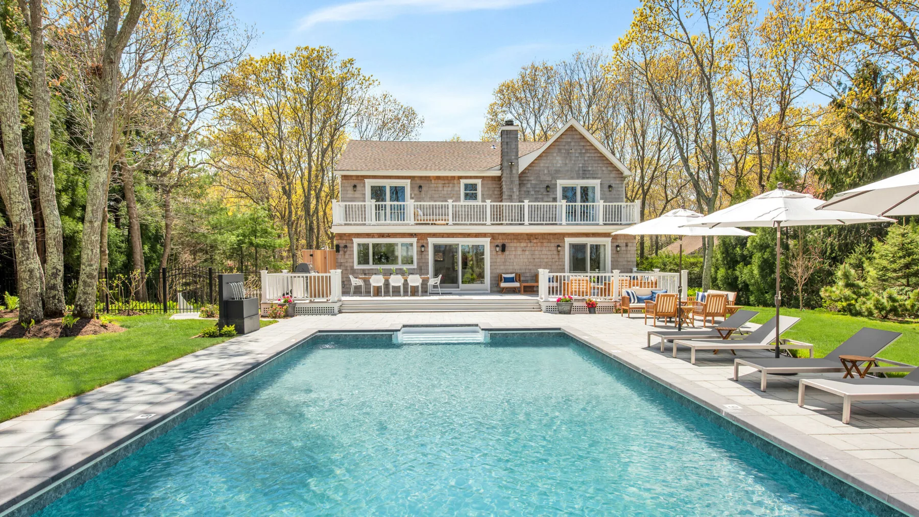 A backyard swimming pool sits in front of a large house, with lounge seating nearby.