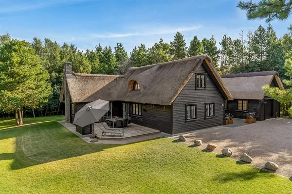 A dark timber house with a thatched roof sits beside a green lawn and trees.