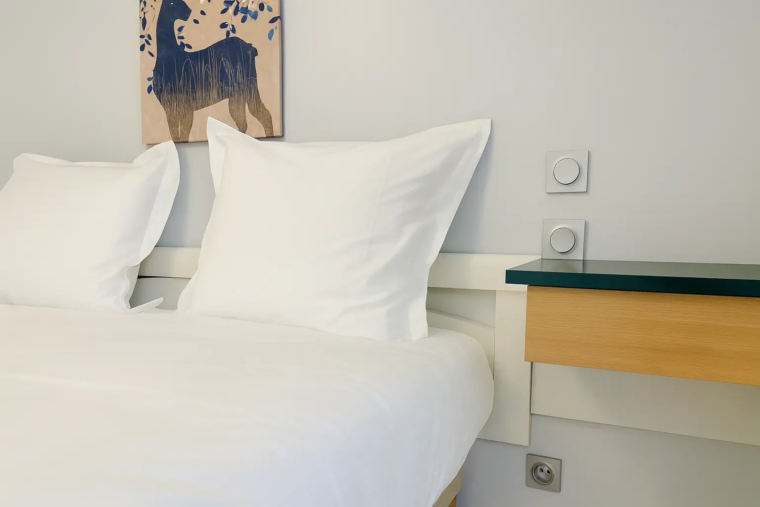 Espacio representativo del hotel: Una cama blanca con almohadas está junto a una mesita de noche y controles en la pared.