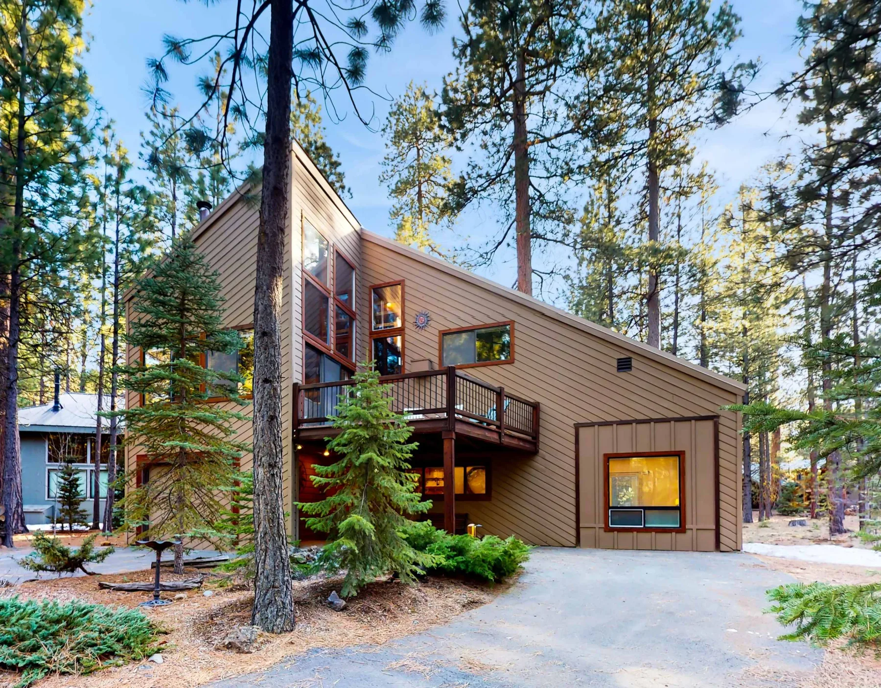 Una casa marrón de forma angular está entre altos pinos, junto a una entrada pavimentada.