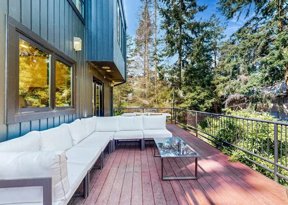 An outdoor deck with white seating and a glass-topped table overlooks leafy trees.
