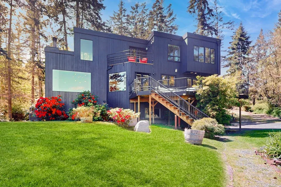 A modern dark exterior house stands among trees beside a green lawn.