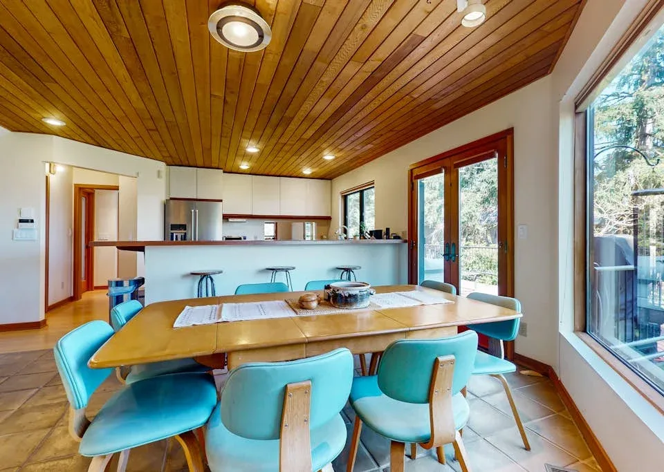 A dining area with a wooden table and turquoise seating beside large windows.