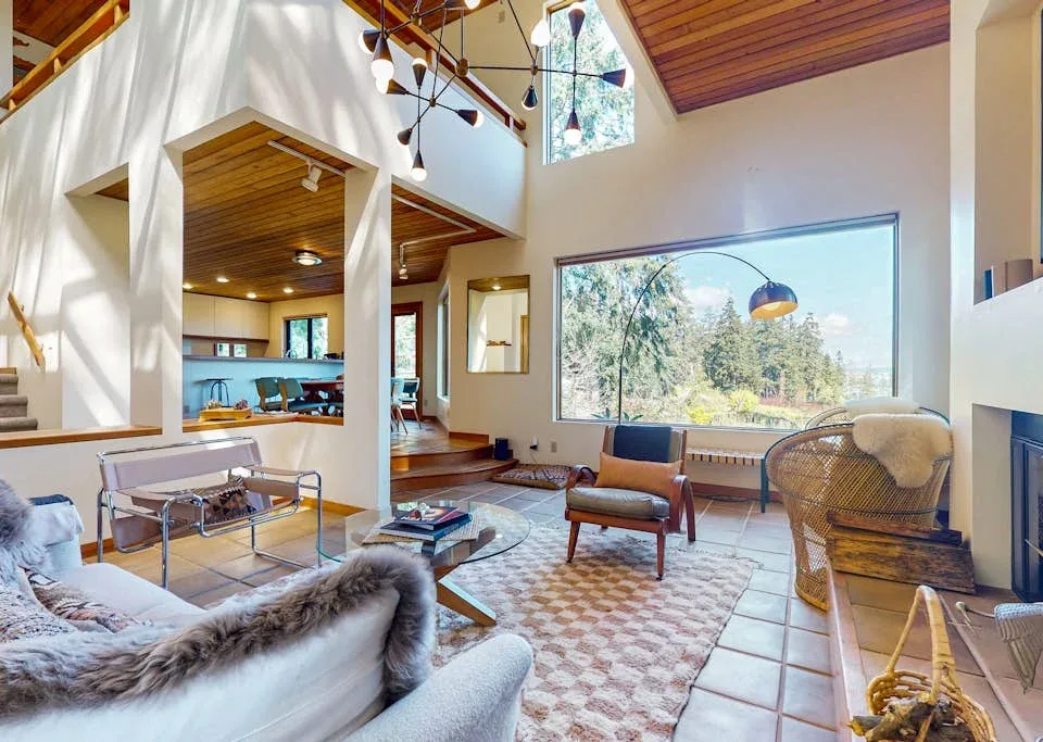 A bright living room with seating, a patterned rug, and a large window overlooking trees.