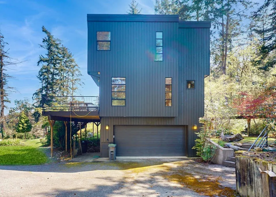 A modern dark-sided house with a garage and raised deck stands among trees.