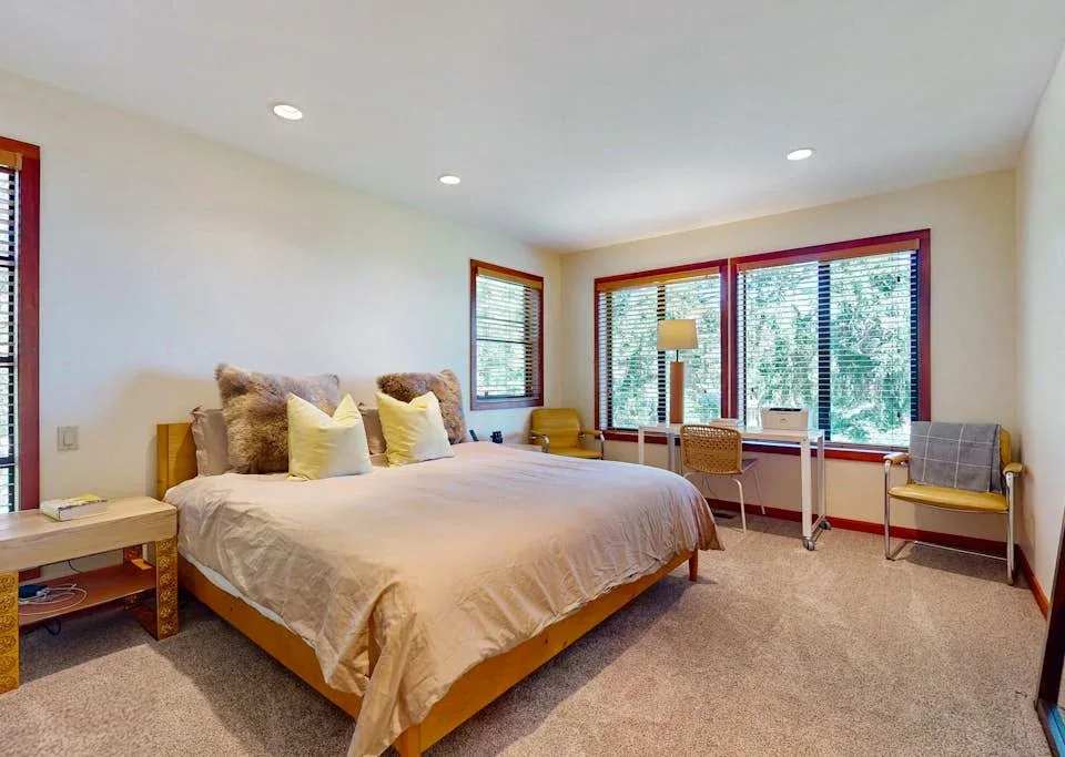 A bedroom with a large bed, desk, and windows overlooking trees.