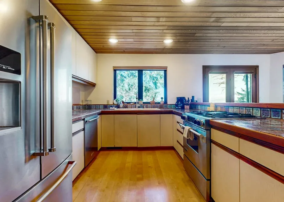 A spacious kitchen with wood cabinetry, stainless steel appliances, and windows.