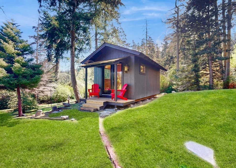 A small cabin sits on a grassy lawn among tall trees, with red chairs outside.