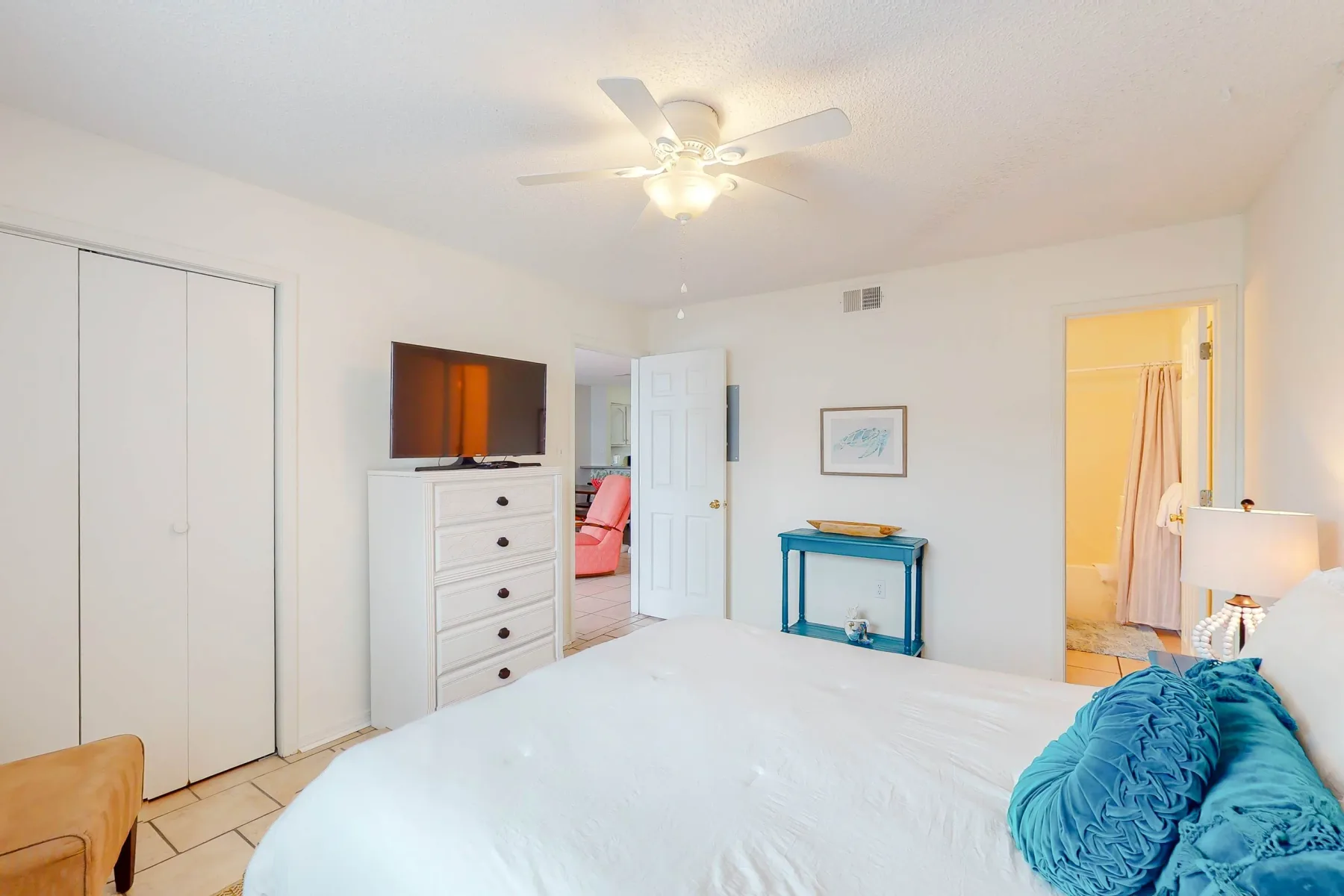 Un dormitorio luminoso con una cama, un ventilador de techo y un televisor sobre una cómoda.