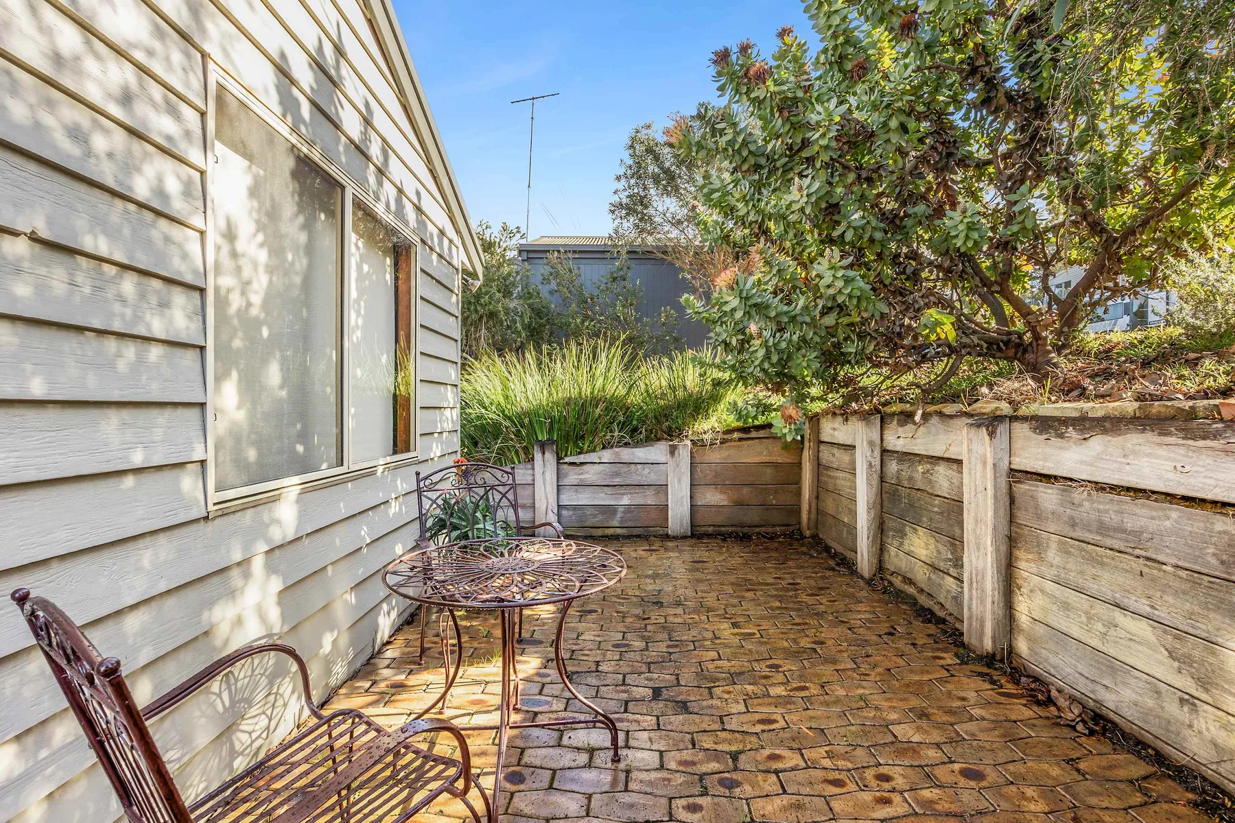 Un patio al aire libre con suelo pavimentado, asientos y un árbol frondoso junto a una casa.