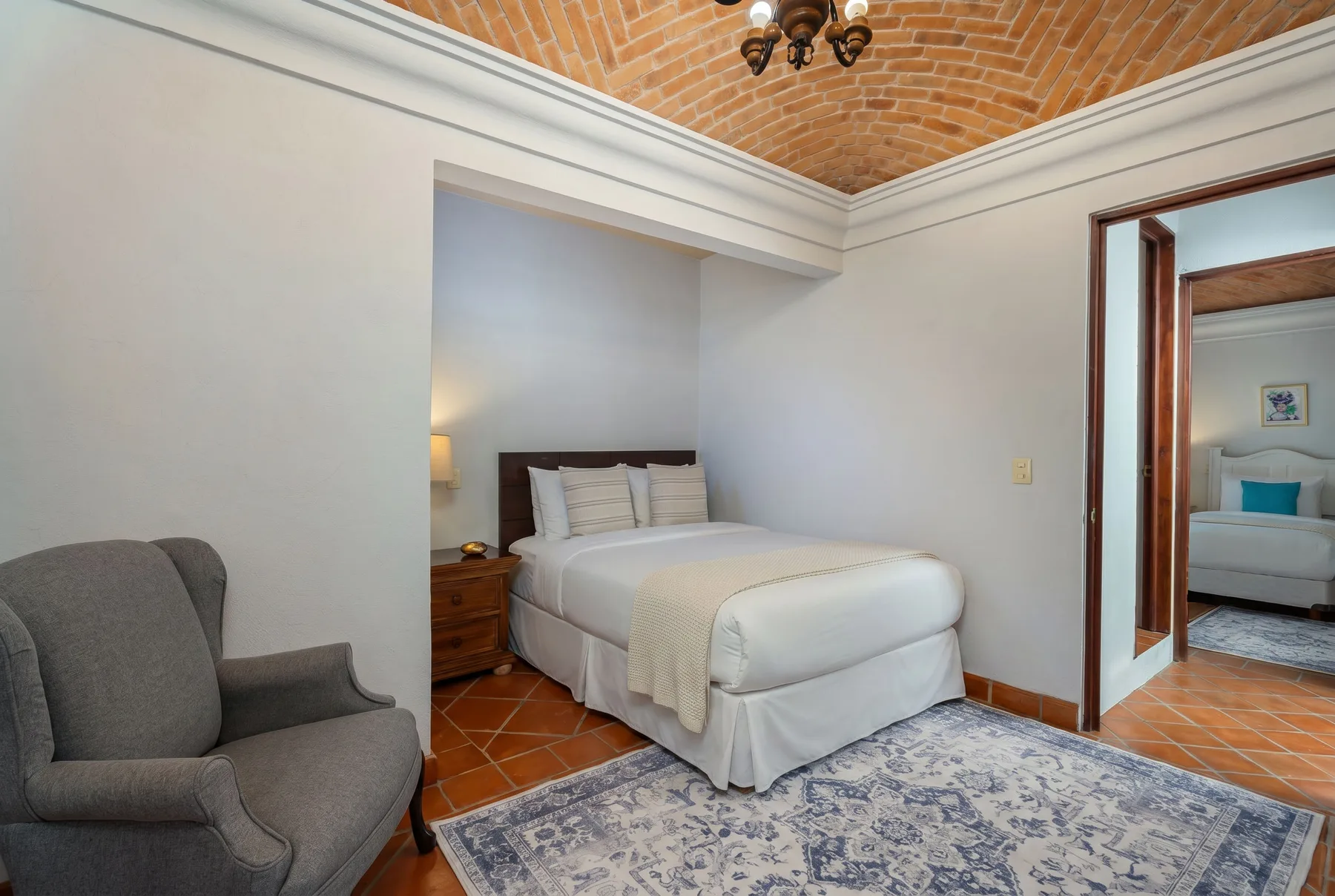 A bedroom with a neatly made bed, an armchair, and a patterned rug.