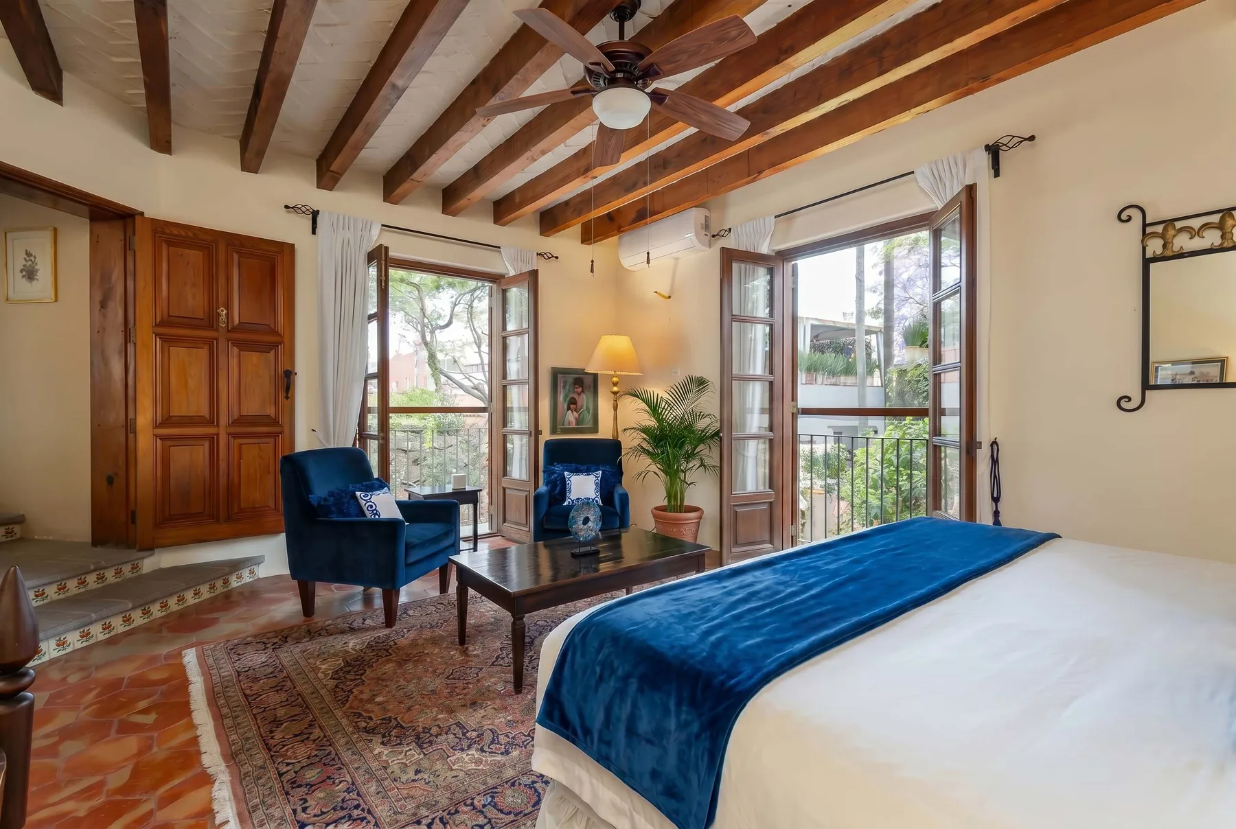 A bedroom with a blue-covered bed, seating, and windows opening onto a balcony.