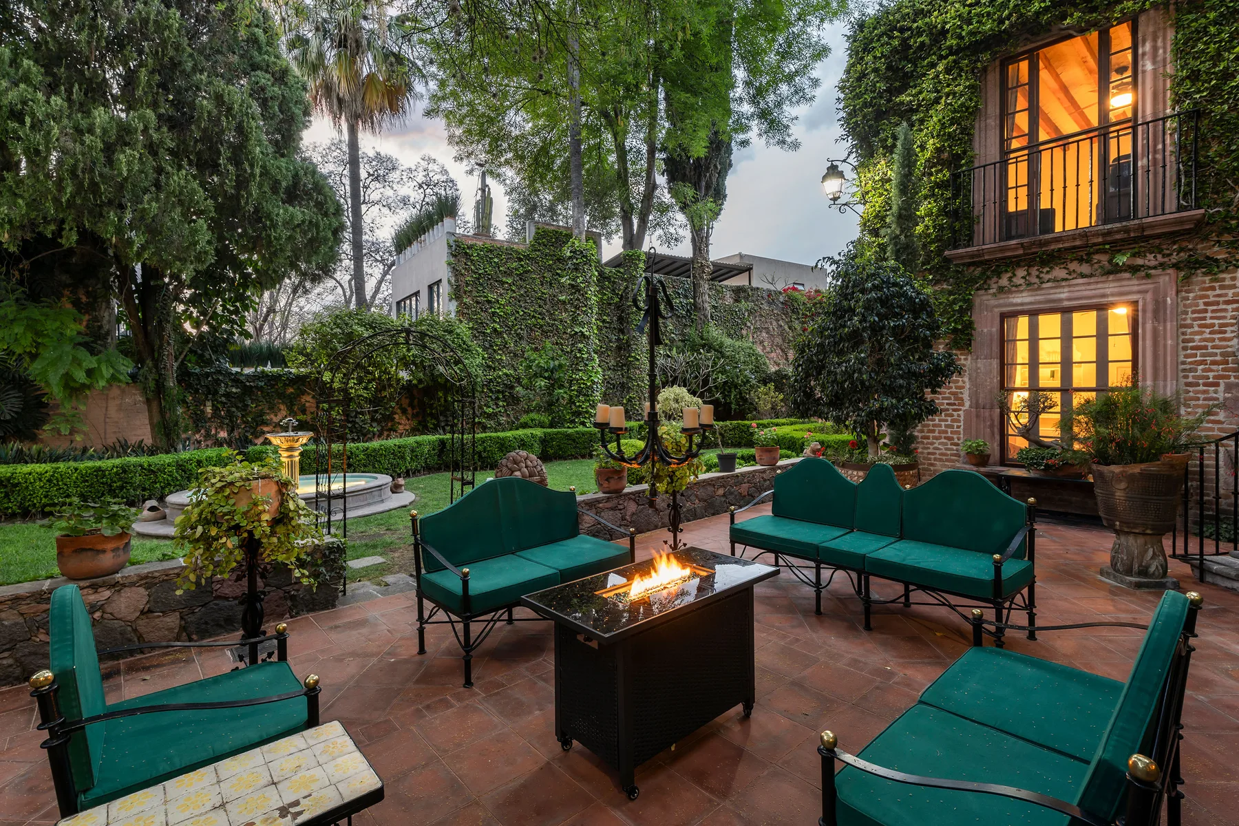 A garden patio has green seating arranged around a lit fire pit.