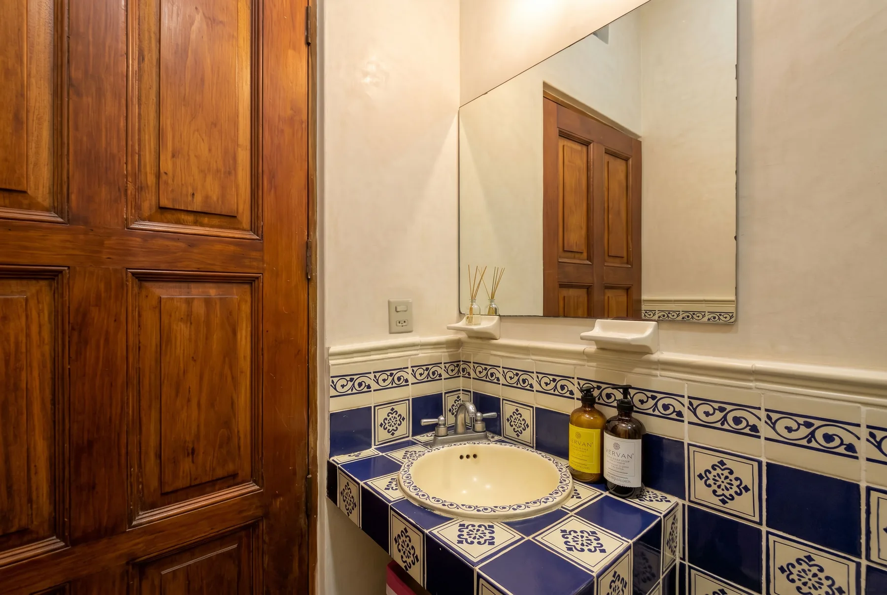 A tiled bathroom vanity with a sink, mirror, and wooden door.