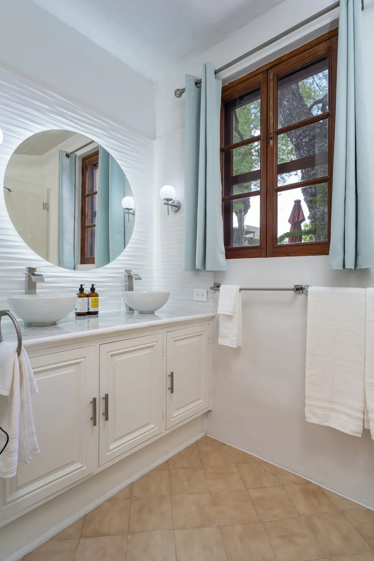 A bright bathroom with a long vanity, round mirror, and wood-framed window.